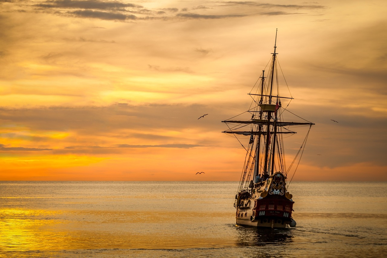 Photo of a large sailing ship moving away from the camera. The sea is calm, the backdrop is of orange clouds.
