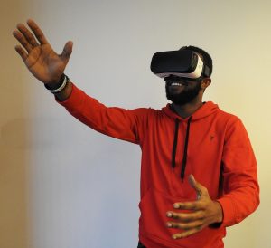 an African American man wearing a VR headset stands in front of a light coloured wall, reaching into the air with his right hand