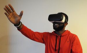 an African American man wearing a VR headset stands in front of a light coloured wall, reaching into the air with his right hand